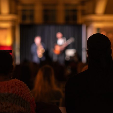 Two performers are out of focus in the distance with the audience in focus and darkness in the foreground.