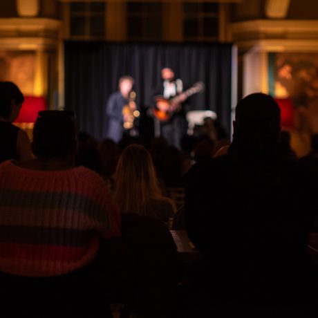 Audience watching a live musical performance with two people with saxophone and guitar on stage.