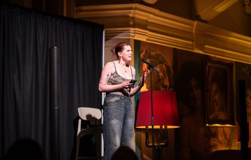 A woman performs on stage speaking into a microphone, illuminated by warm lighting.