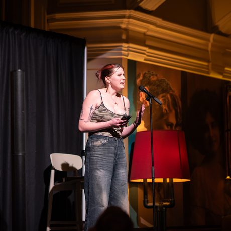 A woman performs on stage speaking into a microphone, illuminated by warm lighting.