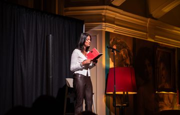 A woman reads from a red book on stage, illuminated by warm lighting.