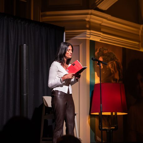 A woman reads from a red book on stage, illuminated by warm lighting.