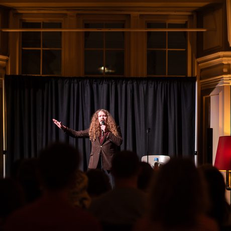 Bradley Taylor on stage in a warmly lit room with attentive audience in the dark.