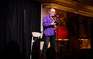 Man speaking on stage with microphone, holding a phone, under warm lighting.