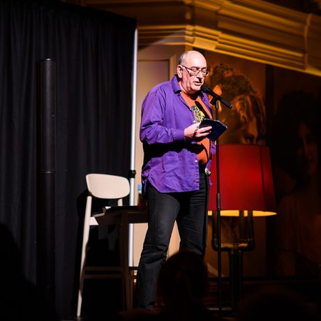 Man speaking on stage with microphone, holding a phone, under warm lighting.