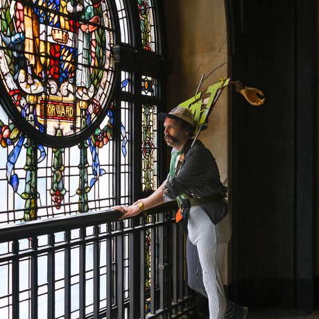 Man with sign that says "TAT" on his shoulders, stands near ornate stained glass window inside building.