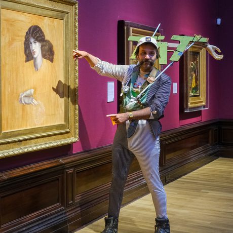 Man dressed in striped suit and large "TAT" sign on his shoulders, pointing at classical painting in art gallery with red walls.