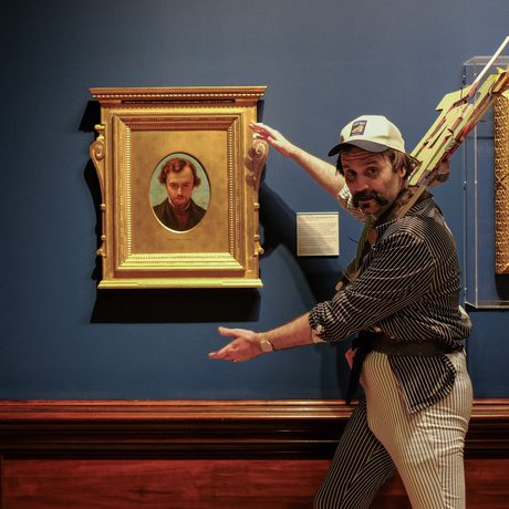 Man in striped suit and cap gestures toward framed portrait in art gallery.