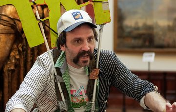 Man wearing a cap and large "TAT" sign, gestures forward. He is indoors in a gallery setting.