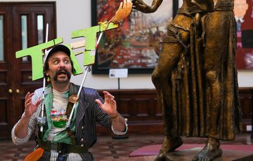 Man in striped suit wears a sign that reads 'TAT' is in a gallery and and interacts playfully with large bronze statue.