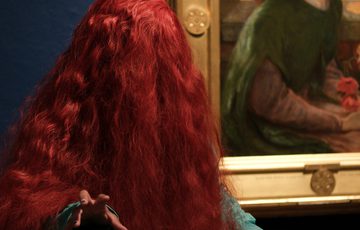 A person in long red wig with their face hidden, in front of a painting of a woman in a holding red flowers.