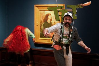 Man with sign that says 'TAT' and person with vibrant red wig perform in front of paintings in an art gallery.