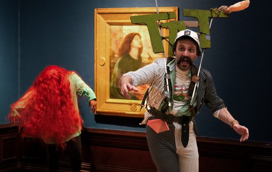 Man with sign that says 'TAT' and person with vibrant red wig perform in front of paintings in an art gallery.