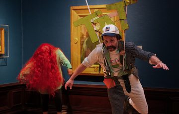 Man with mechanical sign that says 'TAT' and person with vibrant red wig perform in front of paintings in an art gallery.
