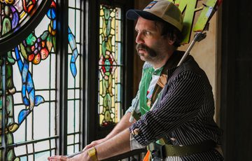 A man wearing a sign that says 'TAT' looks through a colourful stained glass window inside building.