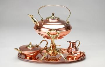 A tea set with teapot, kettle, jug and sugar bowl and tray. Made from shiny copper with accents of brass.