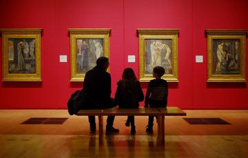 Silhouette of an adult and two children sitting on bench in an exhibition looking at four large paintings in gold frames on the wall in front.