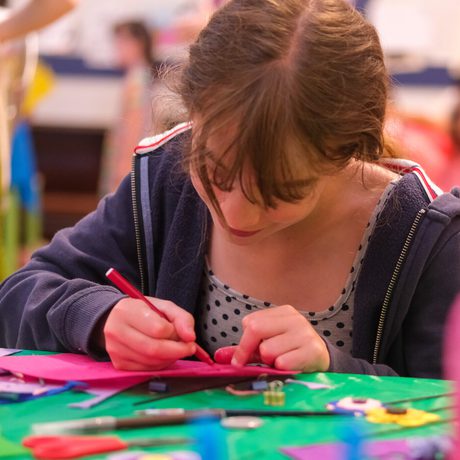 A girl focused on drawing at a colourful, busy table with craft supplies.