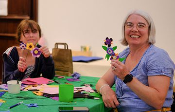Two people smiling, holding colourful handmade felt flowers at a craft table.