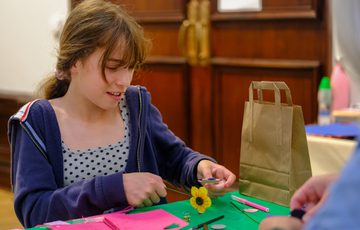 Girl crafting with scissors and paper at a table, next to a paper bag and art supplies.
