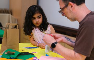 A young girl watches an adult cutting paper for a craft project at a table.