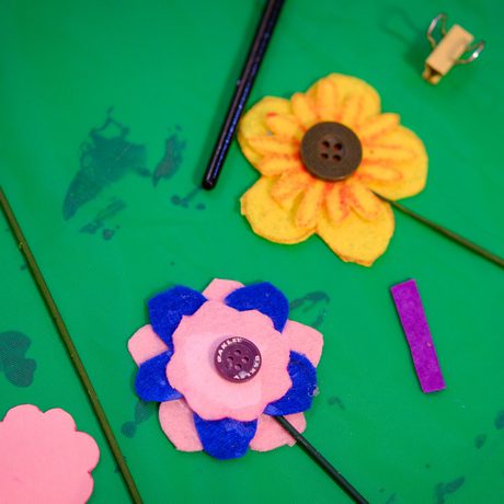 Two handmade fabric flowers with buttons lie on a green surface alongside craft supplies.
