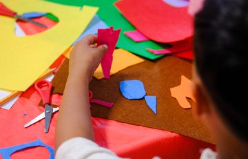 Child crafting colourful paper shapes using scissors on a table covered with crafting materials.