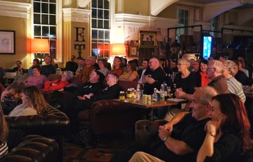 Audience smiling watching a performance or presentation in a warmly lit, cozy room.