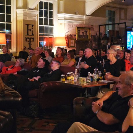 Audience smiling watching a performance or presentation in a warmly lit, cozy room.