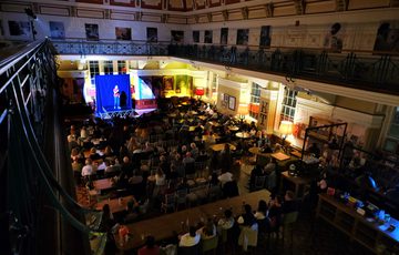 View from a balcony of an audience watching a performance on a small stage inside a large, historic room.