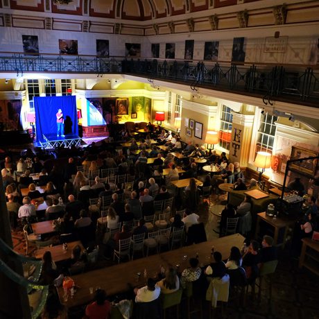View from a balcony of an audience watching a performance on a small stage inside a large, historic room.
