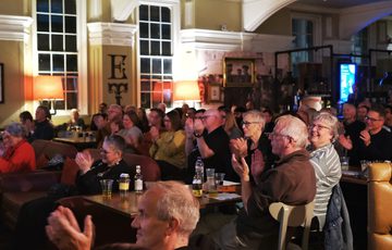 An audience of older adults attentively applauds in a warmly lit event space.