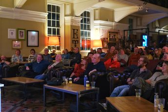 Audience attentively watches a performance or presentation in a cozy, warmly lit lounge.