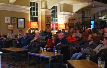 Audience attentively watches a performance or presentation in a cozy, warmly lit lounge.