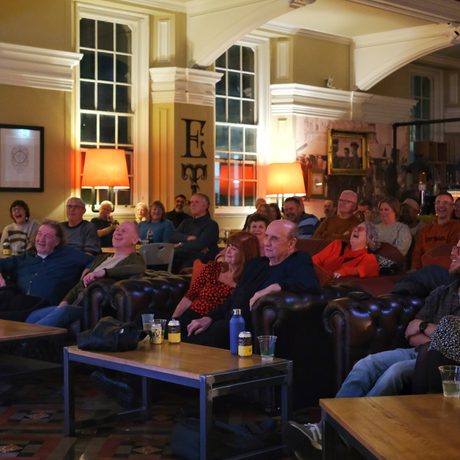 Audience attentively watches a performance or presentation in a cozy, warmly lit lounge.