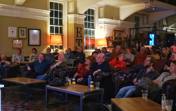 Audience attentively watches a performance or presentation in a cozy, warmly lit lounge.