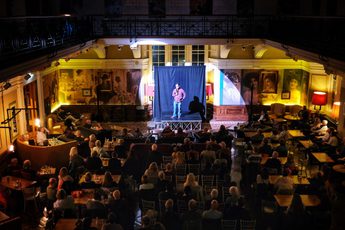 View from a balcony of an audience watching a performance on a small stage inside a large, historic room.