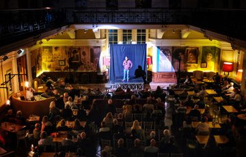 View from a balcony of an audience watching a performance on a small stage inside a large, historic room.