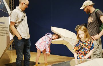 A large 3D model of an life size giant snake with a child putting their head inside and laughing, adults are around them.