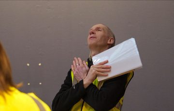 A staff member in high vis jacket standing with arms folded across his chest and looking up while holding paper.