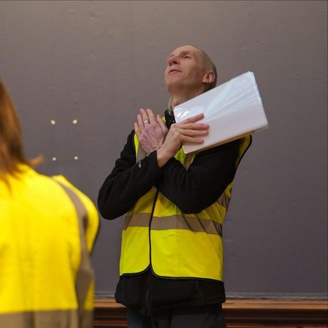 A staff member in high vis jacket standing with arms folded across his chest and looking up while holding paper.