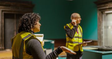 A staff member in a high vis vest with another staff member behind, an empty gallery is behind them.