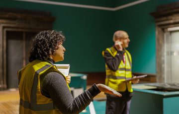 A staff member in a high vis vest with another staff member behind, an empty gallery is behind them.
