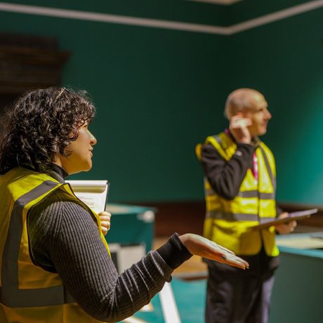 A staff member in a high vis vest with another staff member behind, an empty gallery is behind them.