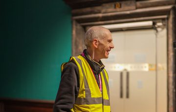 A staff member wearing a high vis vest is smiling in an empty gallery behind.
