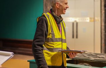 A staff member wearing a high vis vest is smiling in an empty gallery behind.