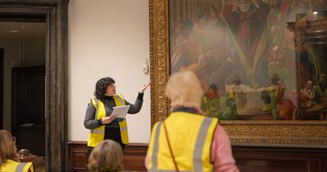 A staff member pointing to a large waiting on display and giving a tour to a group of people wearing high vis vests.
