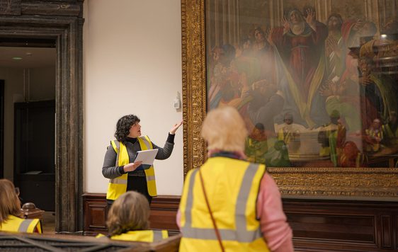 A staff member pointing to a large waiting on display and giving a tour to a group of people wearing high vis vests.