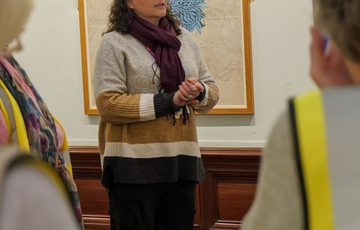 A staff member facing two two people with their backs to the camera. An artwork is on display behind the staff member.