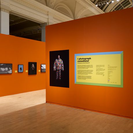 An art gallery exhibition featuring portraits and descriptive panels on vibrant orange walls.